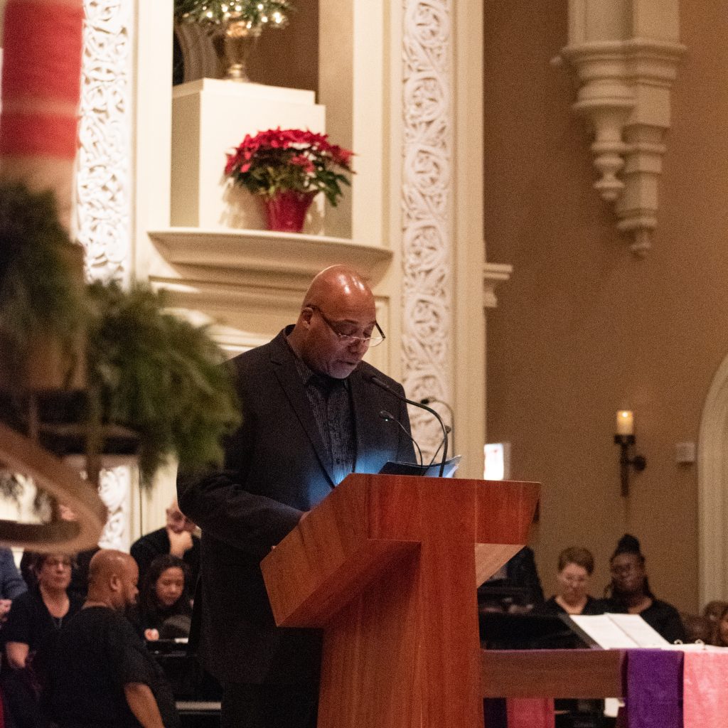 Wayne Richard standing at a at a lectern in a church, surrounded by holiday greenery and with choir members in the background. 