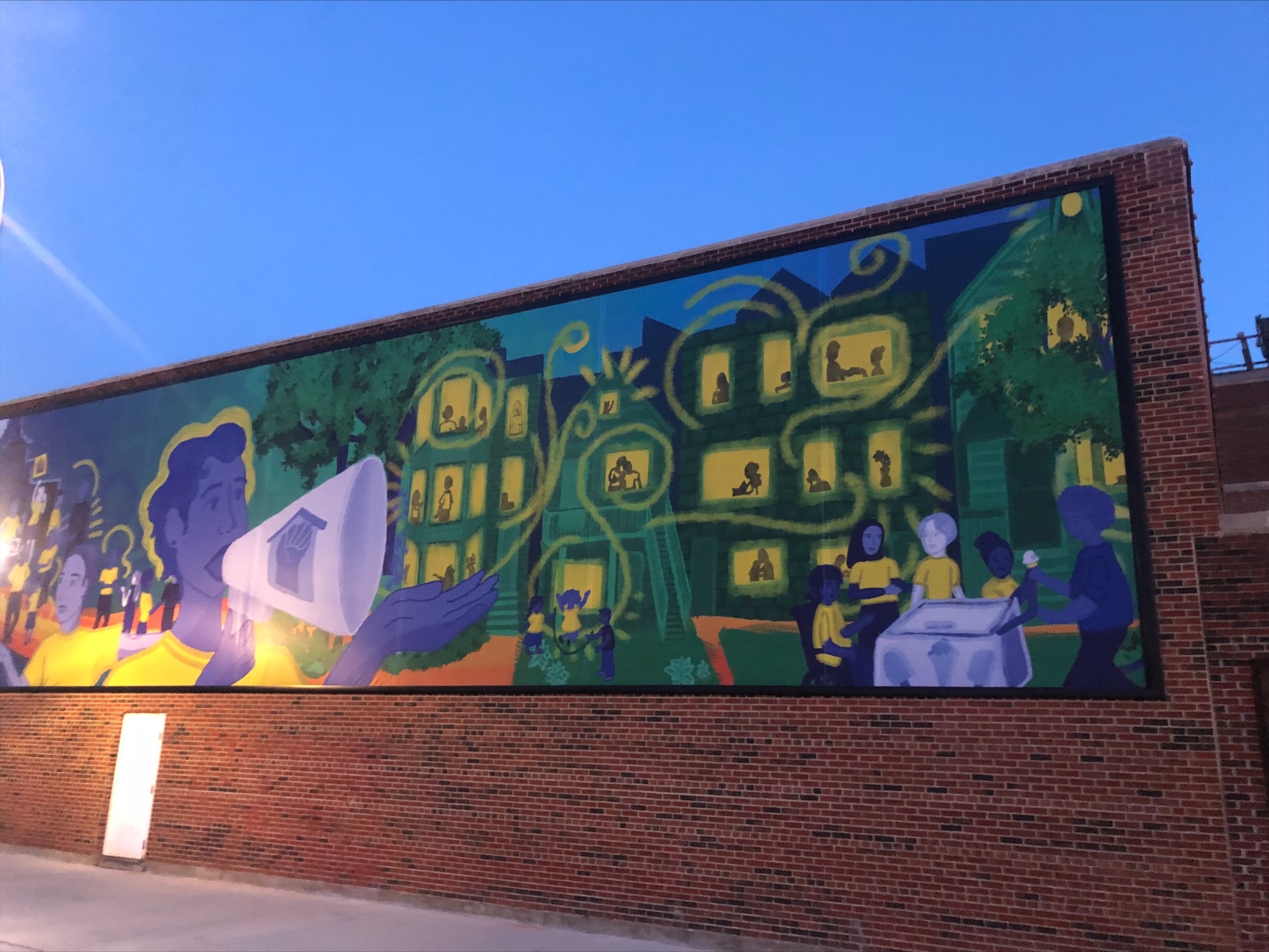 A brick building with a mural on it. The mural features a person with a megaphone speaking to a series of houses whose residents are in their windows. 