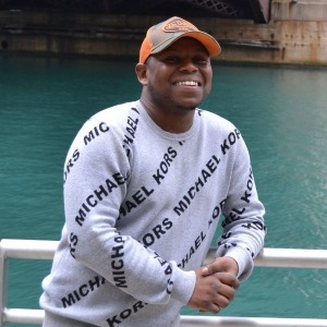 Shamaje, a young Black man, smiles while posing in front of the Chicago River. He is wearing a orange baseball cap and a gray Michael Kors sweater.