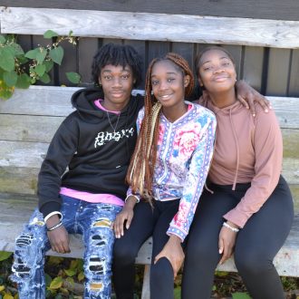 Three Black siblings (ages 13, 14, and 16) pose smiling, sitting on a wooden bench besides a shed.