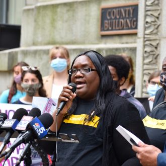 A Black woman speaks to a crowd using a microphone at a press conference in front of Chicago City Hall. She is surrounded by various people of different ages and races, many wearing protective face masks.
