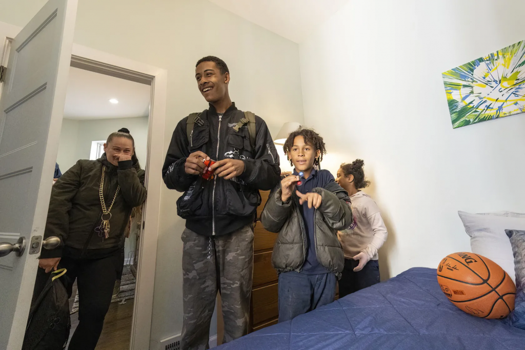 Three kids stand in a bedroom beside a brown dresser and a blue bed with a basketball on it. A mother stands in the doorway, her hand to her face bearing a huge smile. 