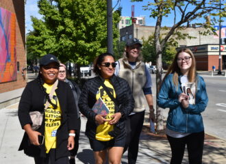 Five young woman of different races walk together on a Chicago sidewalk, smiling. Two women at the front are wearing yellow Chicago Coalition for the Homeless t-shirts.