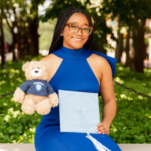 Nia Hill, a young Black woman, smiling. She is seated and wearing a blue dress and tortoise-shell glasses. She is holding a light blue mortarboard and a stuffed bear wearing a Columbia University shirt.
