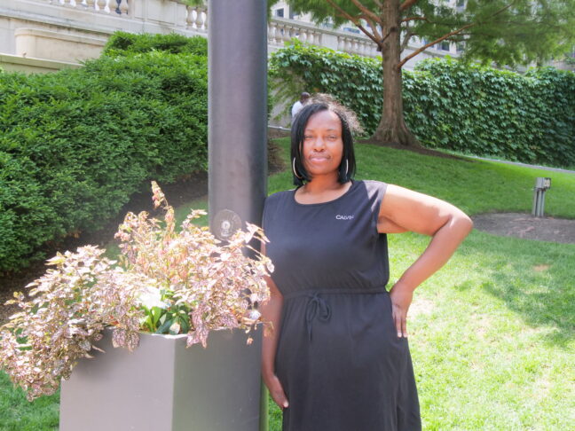Latoya Woffard, a woman in a black dress, leans against a planter.