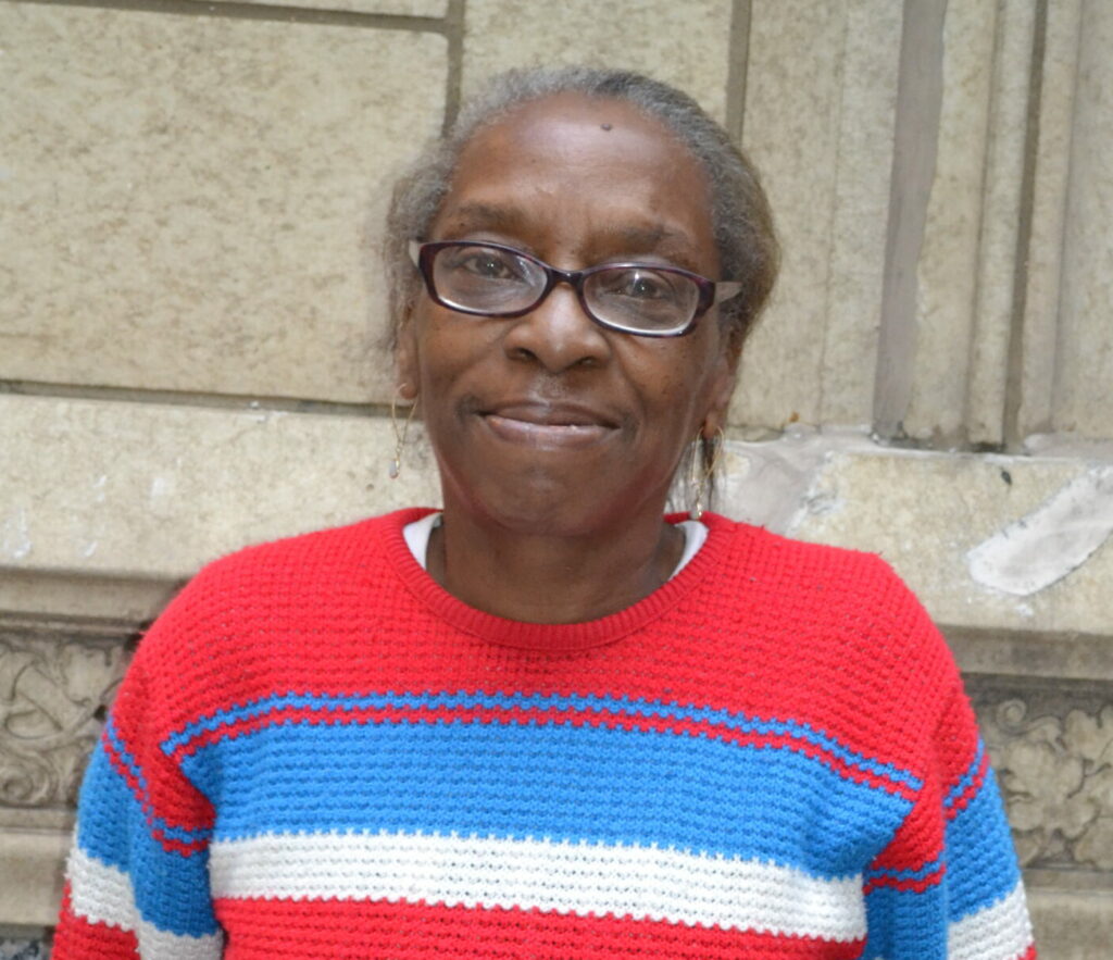 Smiling portrait of a Black woman in her 60s. She is wearing glasses, delicate gold earrings, and a red, white, and blue stripped knit sweater.