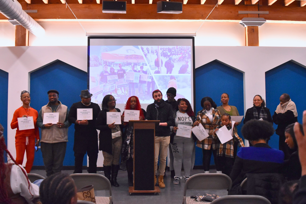 A group of 13 multi-racial, multi-generational people stand side by side behind a podium. Most are holding certificates.