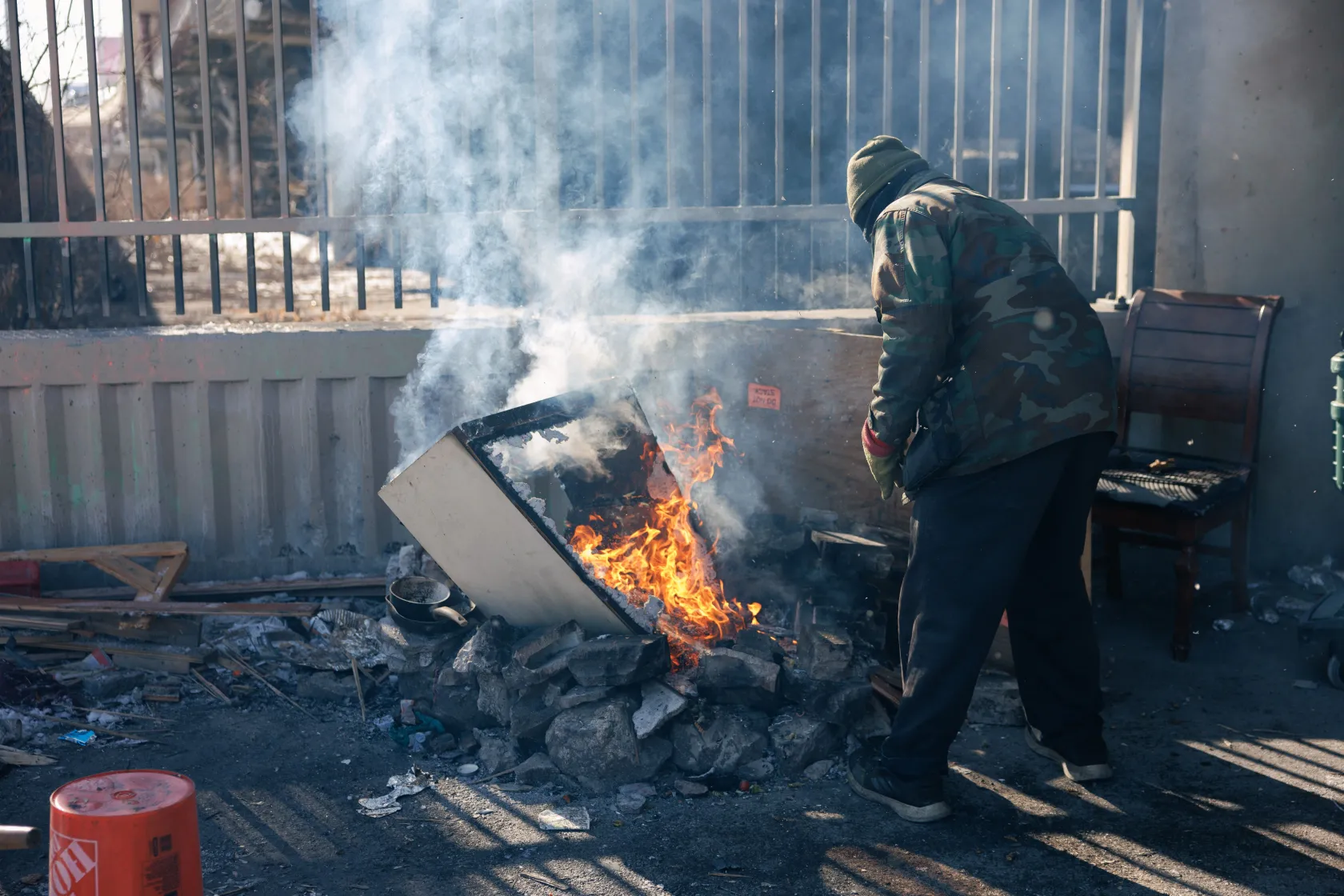 A man in a heavy parka warms himself by a trash fire.