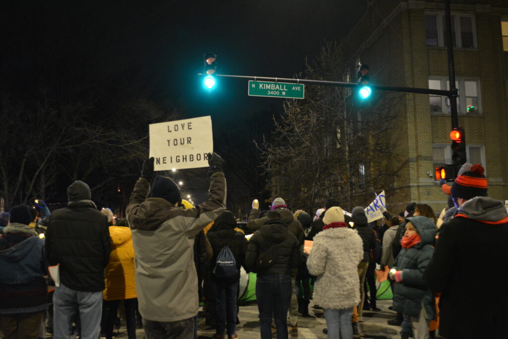 A group of protestors stands at an intersection under a green traffic light. One protestor's sign reads "Love Your Neighbor."