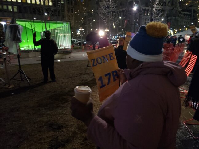 The back of a woman in a beanie holds a yellow sign reading "Zone 17B."