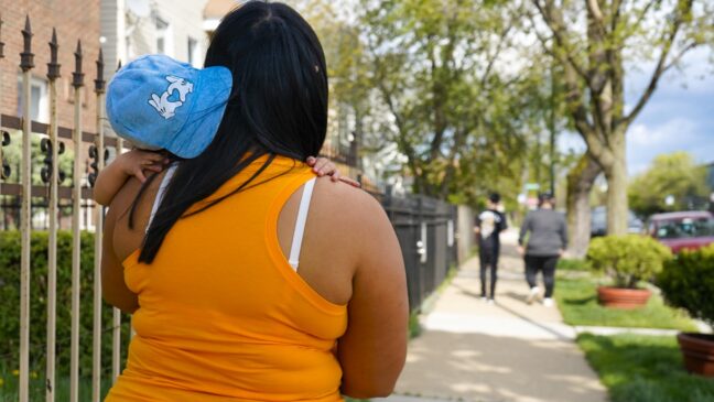Wendy Benitez in an orange shirt and her daughter in a blue cap walk down the sidewalk.